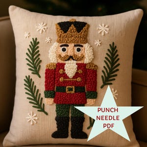Op de afbeelding: Een beige vierkant kussen met een punch needle ontwerp van een notenkraker soldaat. De notenkraker draagt een rode jas, groene broek en zwarte laarzen. Het ontwerp bevat sneeuwvlokken, een kroon en de tekst "PUNCH NEEDLE PDF" in een ster.