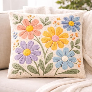 Op de afbeelding: Vierkantig sierkussen met geborduurde bloemen in roze, geel, blauw en paars. De bloemen hebben gele centra en staan tegen een crèmekleurige achtergrond met groene bladeren.