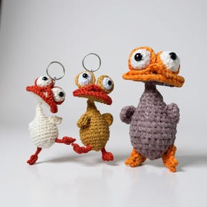 Può includere: Tre portachiavi a forma di anatra fatti a mano all'uncinetto in bianco, giallo e viola. Ogni anatra ha grandi occhi mobili, becchi e zampe arancioni e un portachiavi argentato. Le anatre sono su una superficie bianca.