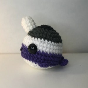 May include: A crocheted plush toy shaped like a narwhal with a white, gray, and purple striped body and a black button eye.
