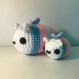 May include: Two crocheted bee plushies, one larger than the other. The larger bee is white with blue and pink stripes, while the smaller bee is white with blue and pink stripes. Both bees have black eyes and a small crocheted wing.