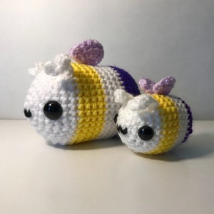 May include: Two crocheted bee plushies, one larger than the other. The larger bee is white, yellow, and purple, while the smaller bee is white, yellow, and lavender. Both bees have black eyes and are smiling.