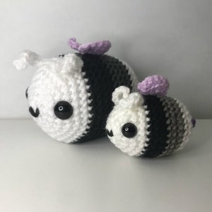 May include: Two crocheted bee plushies, one larger than the other. Both are white and black with purple wings. The larger bee has a black and gray striped body, while the smaller bee has a black and gray striped body with a gray stripe at the bottom.