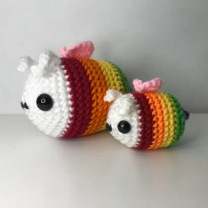 May include: Two crocheted rainbow bee plushies. The larger bee is white with a red, orange, yellow, green, blue, and purple striped body. The smaller bee is the same colors but is smaller in size.