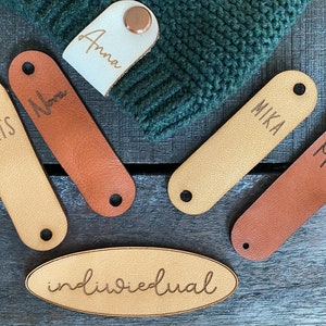 Könnte beinhalten: Fünf personalisierte Leder-Etiketten mit den Namen "Mats", "Nora", "Mika", "Mats" und "Anna". Ein ovales Holzetikett mit dem Wort "individuell" ist ebenfalls zu sehen. Die Etiketten befinden sich auf einer Holzoberfläche.