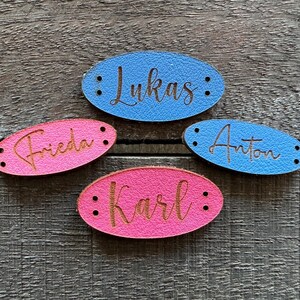 Könnte beinhalten: Vier ovale Lederanhänger in Blau und Pink, jeweils mit einem Namen in Schreibschrift graviert. Die Namen sind Lukas, Frieda, Anton und Karl. Jeder Anhänger hat zwei kleine Löcher an jedem Ende.