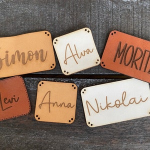Könnte beinhalten: Sechs Namensschilder aus Leder in verschiedenen Braun- und Beigetönen. Jedes Schild trägt einen eingravierten Namen: Simon, Alva, Moritz, Levi, Anna und Nikolai. Die Schilder sind rechteckig oder quadratisch mit abgerundeten Ecken und kleinen Nieten.