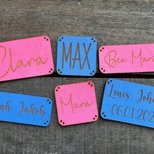 Könnte beinhalten: Sechs personalisierte Leder-Etiketten mit verschiedenen Namen und Daten. Die Etiketten sind in blau und rosa mit schwarzer Schrift.