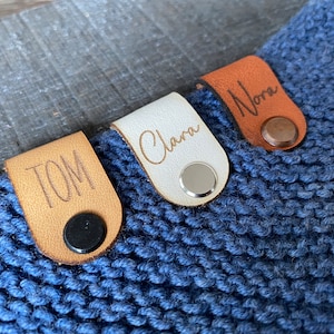 Könnte beinhalten: Drei personalisierte Leder-Namensschilder mit Metallschnappern, die an einem blauen Strickstoff befestigt sind. Die Namen auf den Schildern sind "Tom", "Clara" und "Nora".