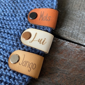 Könnte beinhalten: Drei Leder-Namensschilder mit Metallschnappern. Die Schilder sind braun, weiß und beige. Die Namen "Mats", "Lui" und "Jango" sind auf den Schildern graviert.