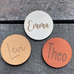 Könnte beinhalten: Drei runde Platzkarte aus Leder mit den Namen "Emma", "Levi" und "Theo" in Kursivschrift graviert.