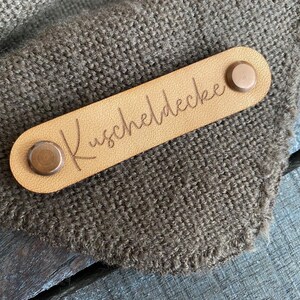 Könnte beinhalten: Ein braunes Leder-Etikett mit zwei Messingnieten und dem Text "Kuscheldecke" in Kursivschrift.