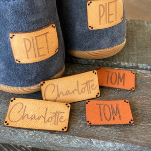 Könnte beinhalten: Vier personalisierte Leder-Etiketten mit den Namen "Piet", "Charlotte" und "Tom" darauf. Die Etiketten sind braun und orange und haben einen genähten Rand.