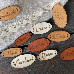 Könnte beinhalten: Zehn ovale Leder-Etiketten mit verschiedenen Namen und einem Vogel-Design. Die Etiketten sind braun, beige und weiß. Die Etiketten sind handgefertigt und haben an den Rändern gelochte Löcher.
