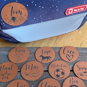 Könnte beinhalten: Zehn braune Leder-Namensschilder mit verschiedenen Designs und Namen, darunter Tom, Teddy, Anja, Coco, Paul, Max, Nikolai, Simon und Bea. Die Namensschilder liegen auf einer Holzoberfläche.