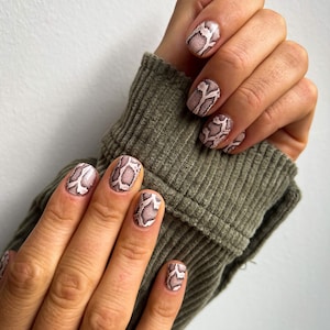 May include: Close-up of a hand with short, oval-shaped nails painted with a snakeskin pattern in shades of brown and pink.