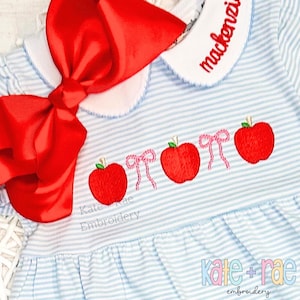May include: A light blue and white striped dress with red embroidered apples and pink bows. A large red bow is attached to the dress. The collar is white with the name "Mackenzie" embroidered in red. The text "Kate Rae Embroidery" is also visible.