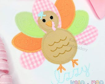 Puede incluir: Un aplique de pavo colorido con cuerpo marrón, pico naranja y plumas multicolores. Las plumas son en tonos de cuadros rosas, naranja y verde. La palabra "lazy" está bordada en escritura azul claro, y el texto "Kate + Rae Embroidery" también es visible.