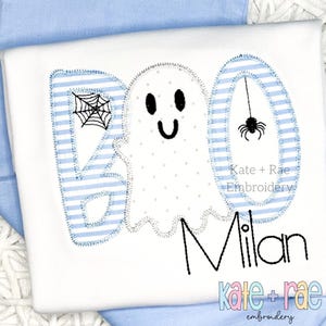 Könnte beinhalten: Eine weiße Applikation mit einem Geist, einem Spinnennetz und einer Spinne, mit dem Wort "BOO" in blau-weißen Streifen und dem Namen "Milan" in Schwarz. Der Text "Kate + Rae Embroidery" ist ebenfalls enthalten.