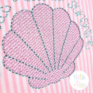 May include: Pink embroidered seashell design with teal stitching on a pink and white striped background. The text "Sunshine" is embroidered in teal. The logo "Kate + Rae" is in the bottom right corner.