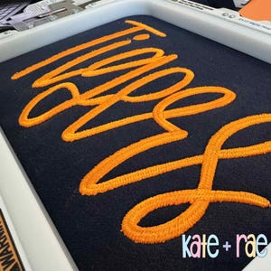 Puede incluir: Primer plano de una tela azul marino con la palabra "Twins" bordada en hilo naranja brillante. El texto est&aacute; en un estilo cursivo. La esquina inferior derecha presenta el texto "kate + rae" en una fuente colorida y estilizada.