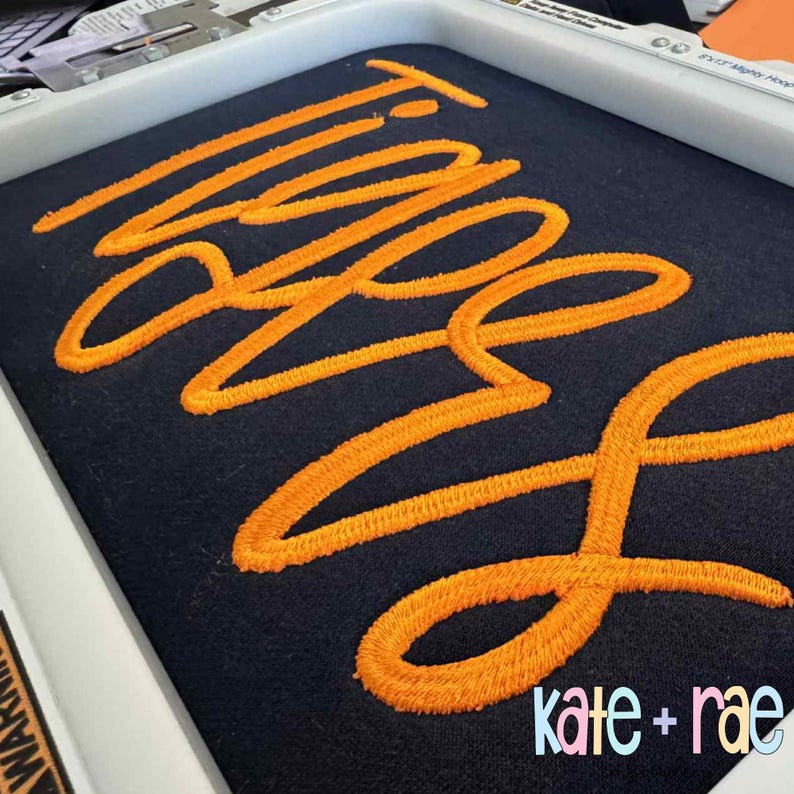 Puede incluir: Primer plano de una tela azul marino con la palabra "Twins" bordada en hilo naranja brillante. El texto est&aacute; en un estilo cursivo. La esquina inferior derecha presenta el texto "kate + rae" en una fuente colorida y estilizada.