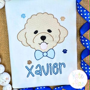 May include: A white t-shirt featuring an embroidered beige dog face with a light blue bow tie, a pink tongue, and small paw prints. The name Xavier is embroidered in blue below the dog. The design includes a watermark reading Kate + Rae.