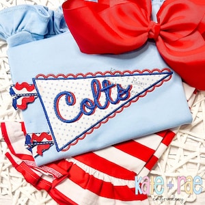 May include: A light blue baby outfit featuring a pennant design with the word "Colts" in blue and red embroidery. The outfit includes a matching red bow and red and white striped bottoms. The brand name "Kate + Rae" is visible.