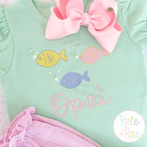 May include: A light green baby onesie with embroidered fish in yellow, pink, and blue. The name "Opal" is embroidered in pink script. A large pink bow is at the top. The onesie is paired with pink and white striped pants.