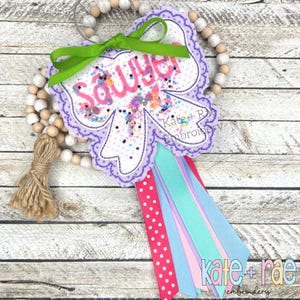 May include: A personalized bow-shaped ornament with the name "Sawyer" in pink. The ornament has a purple scalloped edge, a green bow, and colorful ribbons. The design includes the text "Kate + Rae Embroidery" and is attached to a beaded garland.