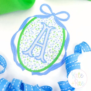 Puede incluir: Un diseño bordado de forma ovalada con un estampado floral y la letra "A". El diseño está delineado en verde y azul, con un lazo de cinta azul en la parte superior. El fondo incluye cintas azules y verdes.