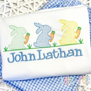 May include: A white babygrow with embroidered blue bunnies holding carrots and the name "John Luathan" in blue thread.