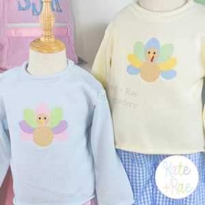 May include: Two long-sleeved knit sweaters, one light blue and one pale yellow, each featuring a central embroidered turkey design with colorful feathers. The text Kate + Rae Embroidery is visible on the yellow sweater and a circular logo.