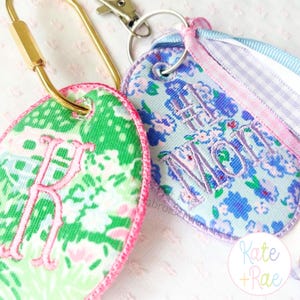 May include: Two embroidered keychains with gold-colored hardware. One keychain has a green floral pattern with a pink embroidered letter "R". The other keychain has a blue floral pattern with the embroidered text "#THE MOM". Both keychains have pink trim.