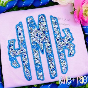 Puede incluir: Una camiseta rosa claro con las letras bordadas "HMH" en un estampado floral con ribete azul. El estampado floral presenta pequeñas flores azules y moradas sobre un fondo blanco. La camiseta está rodeada de cinta azul y vegetación.