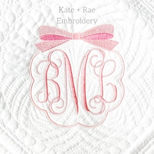 May include: A white fabric with pink embroidery of a monogram "BNE" with a pink bow above it. The text "Kate + Rae Embroidery" is above the monogram.