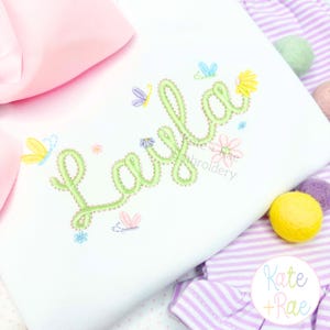 Könnte beinhalten: Weißes T-Shirt mit dem Namen "Layla" in Grün bestickt, umgeben von bunten Schmetterlingen und Blumen. Eine große rosa Schleife ist in der oberen linken Ecke zu sehen. Der Text "Kate + Rae Embroidery" ist ebenfalls vorhanden.