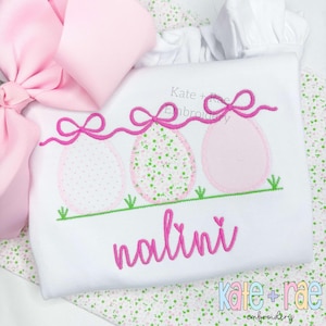May include: A white embroidered shirt with three pink Easter eggs with pink bows and green grass. The name "nalini" is embroidered in pink below the eggs. The shirt is on a white and green floral fabric.