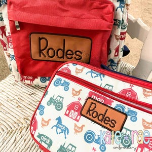 Puede incluir: Una mochila roja y blanca y un estuche a juego con un tema de animales de granja. La mochila tiene un bolsillo frontal rojo con un parche de cuero marrón que dice "Rodes". El estuche tiene una cremallera y un parche de cuero marrón que dice "Rodes".