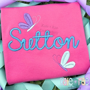 May include: A pink t-shirt featuring the name "Sutton" in turquoise script, with two embroidered butterflies. The butterflies have purple and white wings. The text "Kate + Rae Embroidery" is also visible.