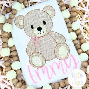 May include: White t-shirt featuring an embroidered teddy bear with a pink bow and the name "Emmy" in pink script. The bear is light brown with a darker brown outline. The shirt is surrounded by wooden beads.