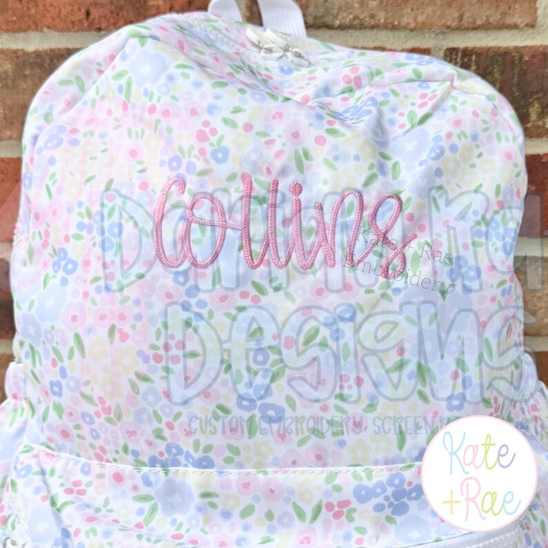 May include: A floral print backpack with the name "Collins" embroidered in pink script. The backpack features a pattern of small pink, blue, and yellow flowers on a white background. The words "Designs" and "Custom Embroidery, Screen" are also visible.