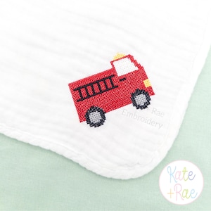 May include: White baby blanket with a red cross-stitch fire truck design. The fire truck has black details and gray wheels. The text "Rae Embroidery" is visible on the blanket.