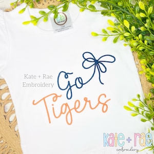 Op de afbeelding: Een witte baby romper met blauwe en oranje borduurwerk met de tekst "Go Tigers" met een blauwe strik boven de tekst. De tekst "Kate + Rae Embroidery" is onder het ontwerp gedrukt.