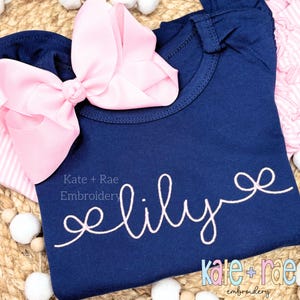 Puede incluir: Una camiseta azul marino con bordado rosa que dice "lily" con un lazo rosa en la parte superior. El texto "Kate + Rae Embroidery" también es visible en la camiseta.
