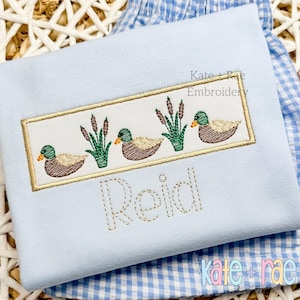 May include: A light blue embroidered shirt with a gold frame featuring three ducks and cattails. The name "Reid" is embroidered below the frame in a gold thread.