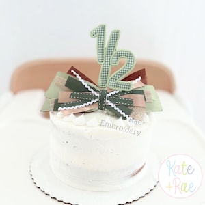 May include: A white frosted cake topped with a decorative bow and a "1/2" cake topper. The bow features layers of green, brown, and tan ribbons. The "1/2" is green gingham. The cake sits on a white cake board. The text "Kate Rae Embroidery" is visible.