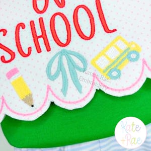 May include: A white fabric piece with a scalloped edge featuring red embroidered text that reads BACK TO SCHOOL. The design includes a yellow pencil, a light blue bow, and a yellow school bus. The fabric has a light pink polka dot pattern.