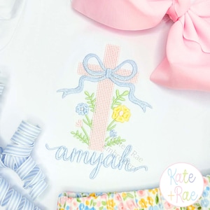 May include: White onesie with a pink cross design, adorned with a light blue bow and floral embroidery. The name "amyah" is embroidered in a matching light blue script. A pink bow and floral patterned shorts are also visible.
