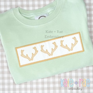 May include: A light green t-shirt with a white rectangle embroidered with three gold antlers. The text "Kate + Rae Embroidery" is printed above the rectangle.